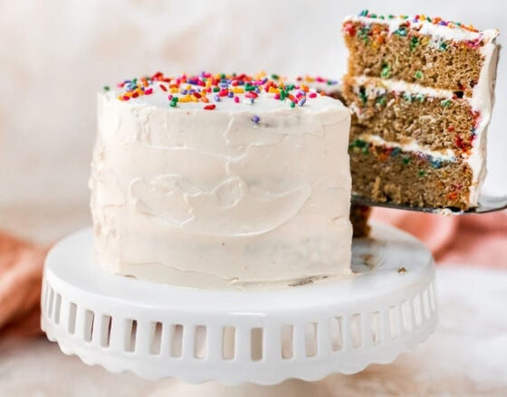 A cake sitting on a cake stand. The cake is iced in white frosting and topped with rainbow sprinkles. A slice has been cut and is being removed, exposing the 3 layers of funfetti cake with cream cheese frosting in between the layers.