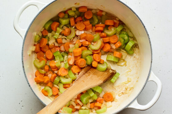 Looking into a dutch oven with chopped and diced celery, carrots and onion. A wooden spoon is pushing the vegetables around the pot.