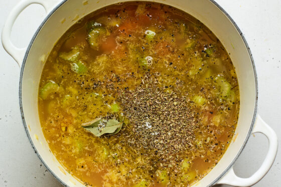 A dutch oven containing chicken broth, bay leaves, thyme, oregano, pepper, salt, carrots, onions, garlic and celery.