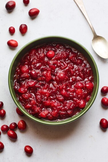 A serving dish containing cranberry sauce. A spoon rests at the top of the bowl and there are whole cranberries scattered around the bowl.