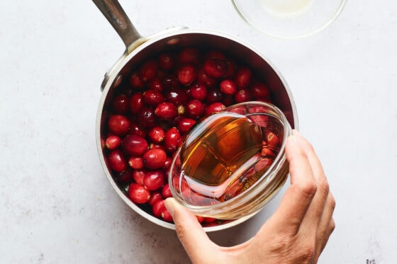 A saucepan containing whole cranberries. A hand is pouring maple syrup into the saucepan from a small ramekin.