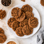 An overhead photo looking down at a plate of almond butter espresso cookies. Extra cookies are scattered around the plate alongside a bowl of coffee beans and a mug of hot cocoa.