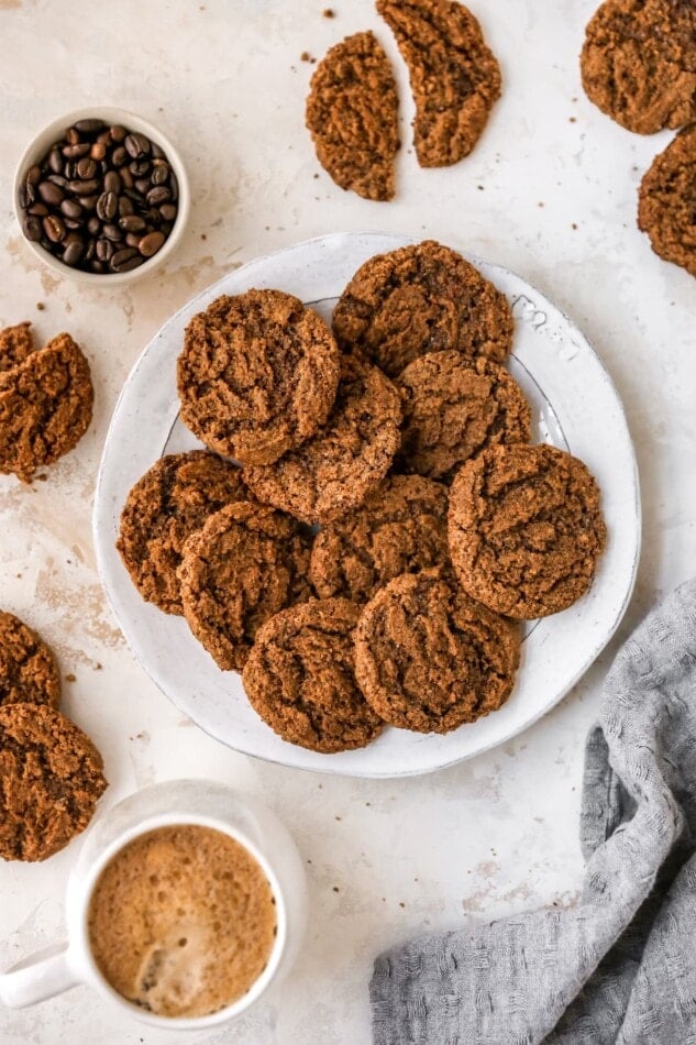 The BEST almond butter espresso cookies! Only six ingredients. Plus, they're flourless, vegan and gluten-free! An overhead photo looking down at a plate of almond butter espresso cookies. Extra cookies are scattered around the plate alongside a bowl of coffee beans and a mug of hot cocoa.