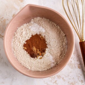 A mixing bowl containing flours, baking powder, baking soda, salt, cinnamon, ground ginger and cloves. A whisk lays next to the bowl.