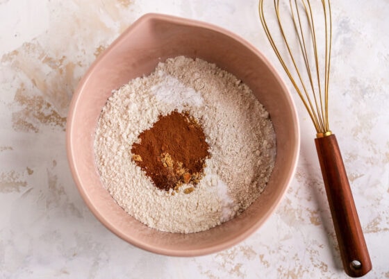 A mixing bowl containing flours, baking powder, baking soda, salt, cinnamon, ground ginger and cloves. A whisk lays next to the bowl.