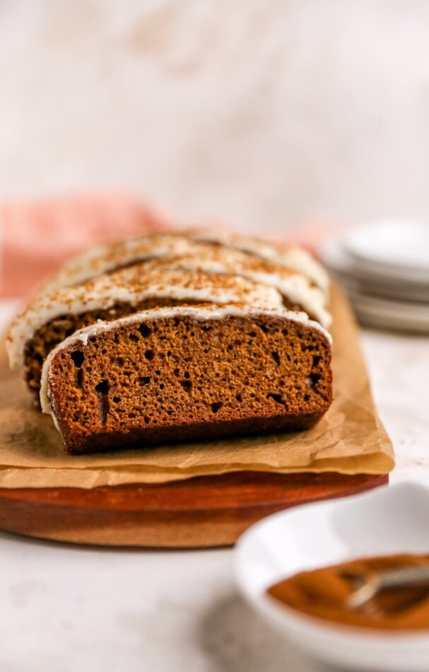 Gingerbread loaf with maple cream cheese frosting sliced on a piece of parchment paper covering a wooden cutting board.