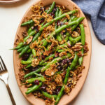 An overhead photo looking down onto a serving dish containing green bean salad topped with fried shallots. A fork is resting next to the serving dish, as well as a blue linen dishcloth.