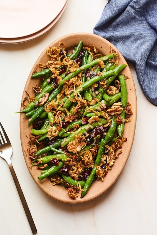 An overhead photo looking down onto a serving dish containing green bean salad topped with fried shallots. A fork is resting next to the serving dish, as well as a blue linen dishcloth.