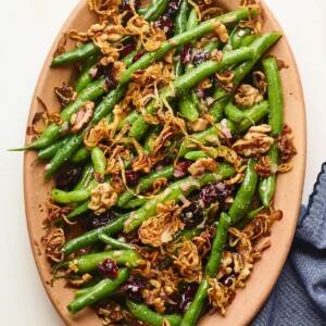 An overhead photo looking down onto a serving dish containing green bean salad topped with fried shallots. A blue linen dish cloth is laying next to the serving dish.