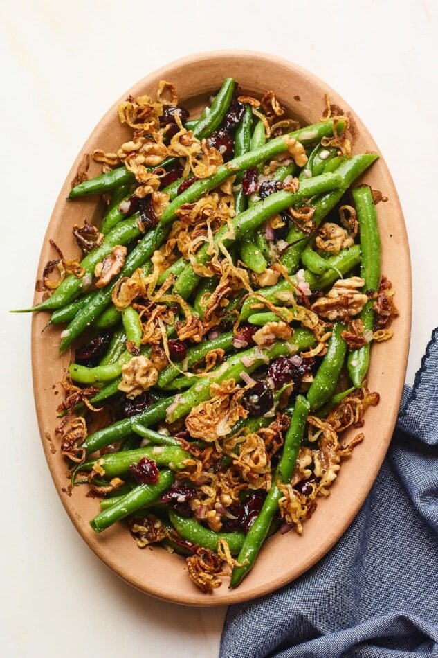 An overhead photo looking down onto a serving dish containing green bean salad topped with fried shallots. A blue linen dish cloth is laying next to the serving dish.