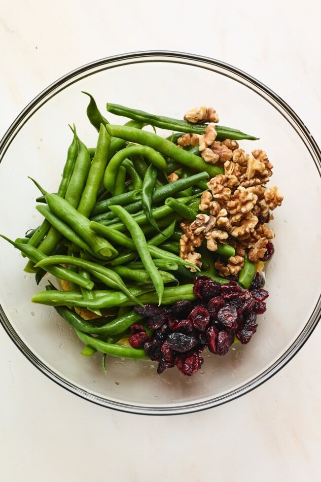 A glass mixing bowl containing blanched green beans, nuts and dried cranberries.