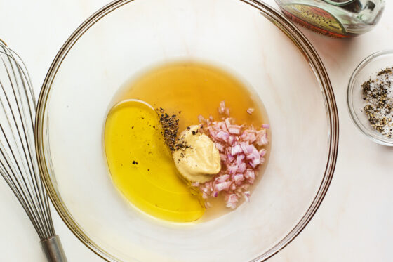 A glass mixing bowl containing apple cider vinegar, olive oil, maple syrup, minced shallots, dijon mustard, sea salt and pepper.