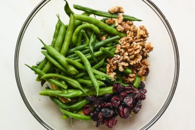 A glass mixing bowl containing blanched green beans, nuts and dried cranberries.