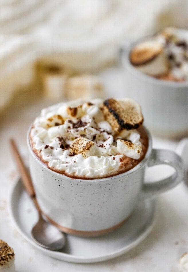 A mug of healthy hot cocoa. It has been topped with whipped cream and toasted marshmallows. A spoon rests next to the mug.