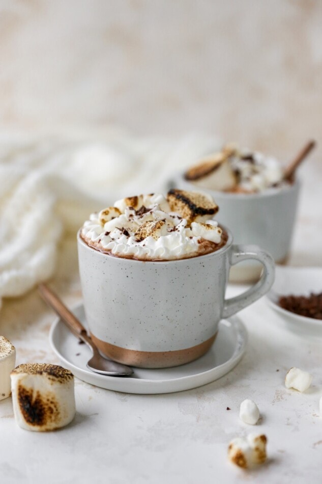 A mug containing healthy hot cocoa, topped with whipped cream and toasted marshmallows. There are marshmallows scattered around the mug and a spoon resting on a plate next to the mug.