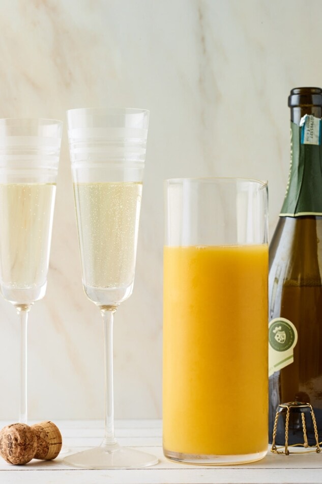 Two Champagne flutes, a bottle of Champagne and a carafe of orange juice. The Champagne flutes have been filled two-thirds of the way with Champagne.
