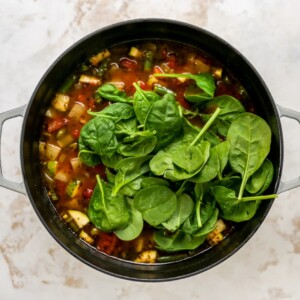 Spinach added to a large pot containing ingredients for minestrone soup.