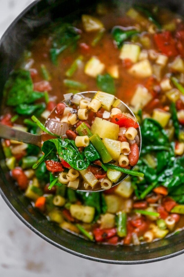 An overhead photo looking down into a large pot of minestrone soup. A ladle is holding up a scoop of the soup close to the lense.