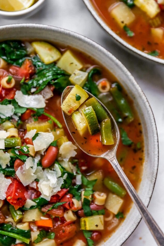 A bowl of minestrone soup. A spoon is holding up a bite of soup.