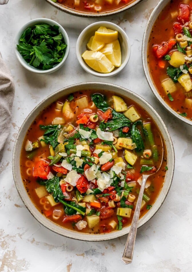 An overhead photo looking down at a bowl of minestrone soup. A spoon rests in the bowl and there are two small bowls with lemon wedges and fresh herbs next to the bowl of soup.