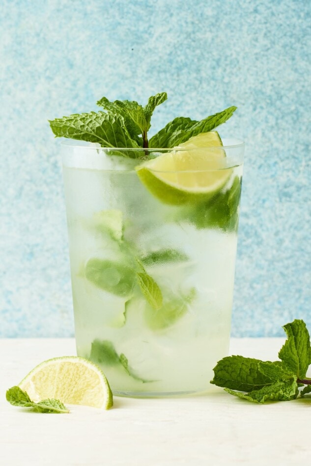 A clear glass containing a skinny mojito. There is a sprig of mint inside the glass alongside of a lime wedge. Extra mint leaves and lime are around the glass on the counter.