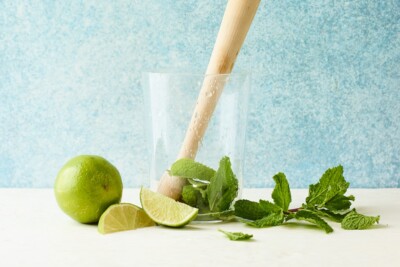 A glass with mint leaves at the bottom being muddled with lime juice by a wooden muddler.