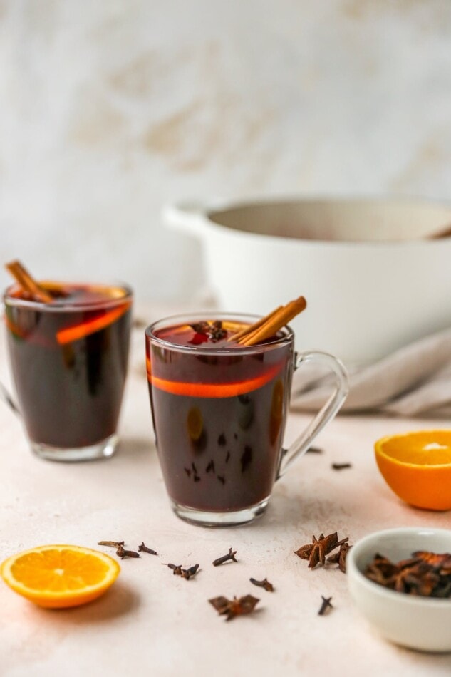 Two mugs of mulled wine. Each mug is topped with orange slices and cinnamon sticks. Extra orange slices, cloves and star anise lay scattered around the mugs.