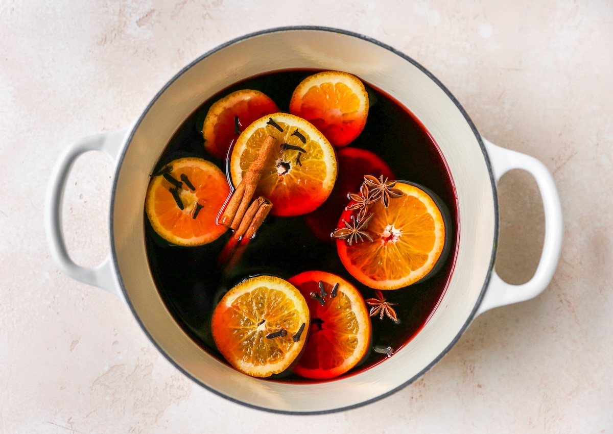 A large pot containing ingredients for mulled wine: red wine, brandy, maple syrup, orange slices, cinnamon sticks, cloves and star anise.