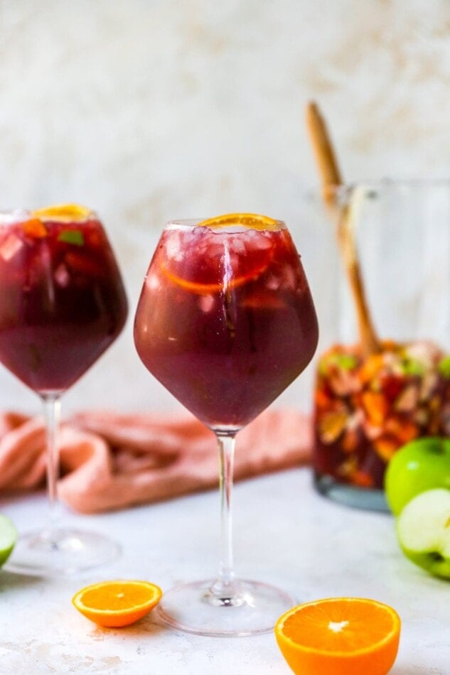 A wine glass containing red wine sangria. There is a second glass out of focus in the background along with a pitcher.