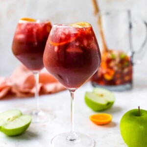 A wine glass containing red wine sangria. There is a second glass out of focus in the background along with a pitcher.