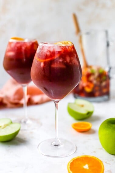 A wine glass containing red wine sangria. There is a second glass out of focus in the background along with a pitcher.