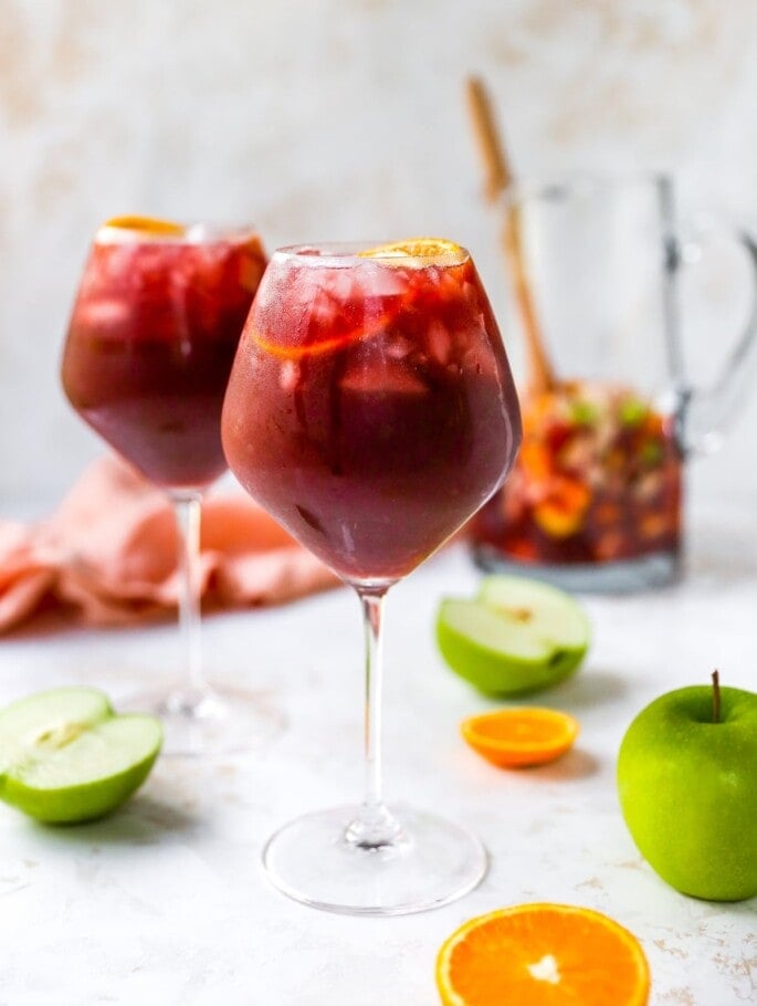 A wine glass containing red wine sangria. There is a second glass out of focus in the background along with a pitcher.