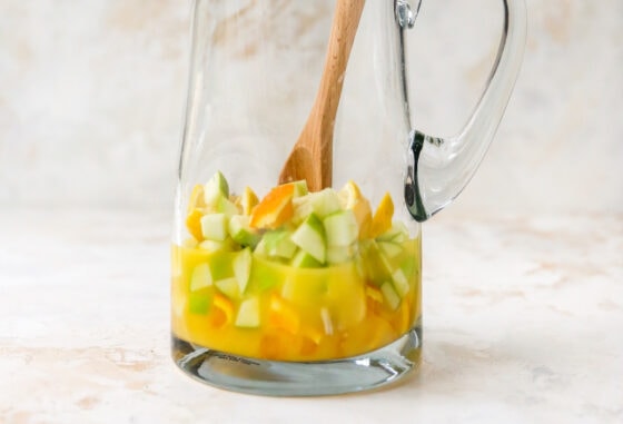 A glass pitcher containing pieces of chopped apple, orange and lemon in orange juice.