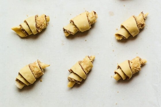 Rugelach wedges rolled into crescent shapes, resting on a sheet pan lined with a piece of parchment paper.