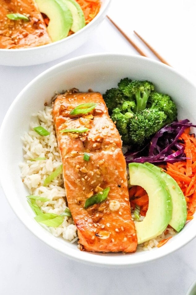 An overhead photo looking at a bowl of rice topped with a filet of teriyaki salmon, broccoli, shredded carrots, shredded cabbage, green onion and sliced avocado.