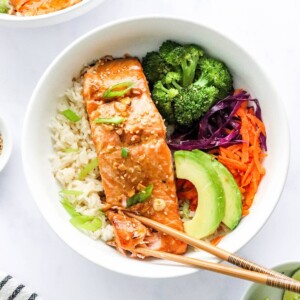 An overhead photo looking at a bowl of rice topped with a filet of teriyaki salmon, broccoli, shredded carrots, shredded cabbage, green onion and sliced avocado. There are two chopsticks resting on the bowl.