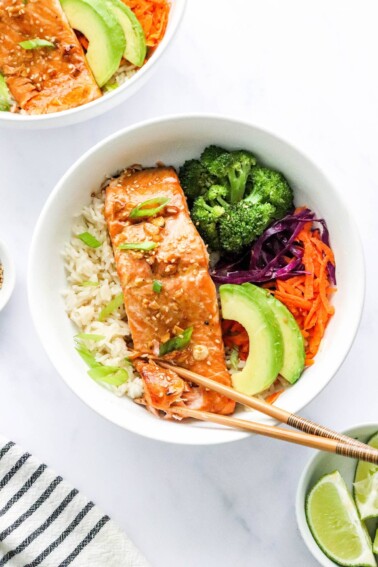 An overhead photo looking at a bowl of rice topped with a filet of teriyaki salmon, broccoli, shredded carrots, shredded cabbage, green onion and sliced avocado. There are two chopsticks resting on the bowl.