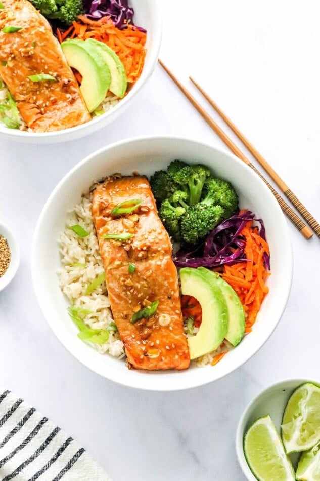 An overhead photo looking at a bowl of rice topped with a filet of teriyaki salmon, broccoli, shredded carrots, shredded cabbage, green onion and sliced avocado. There are chopsticks resting next to the bowl alongside another bowl just out of frame.
