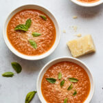 Three bowls containing tomato basil soup. Each bowl is topped with fresh basil leaves and there is a wedge of parmesan cheese resting near the bowls.