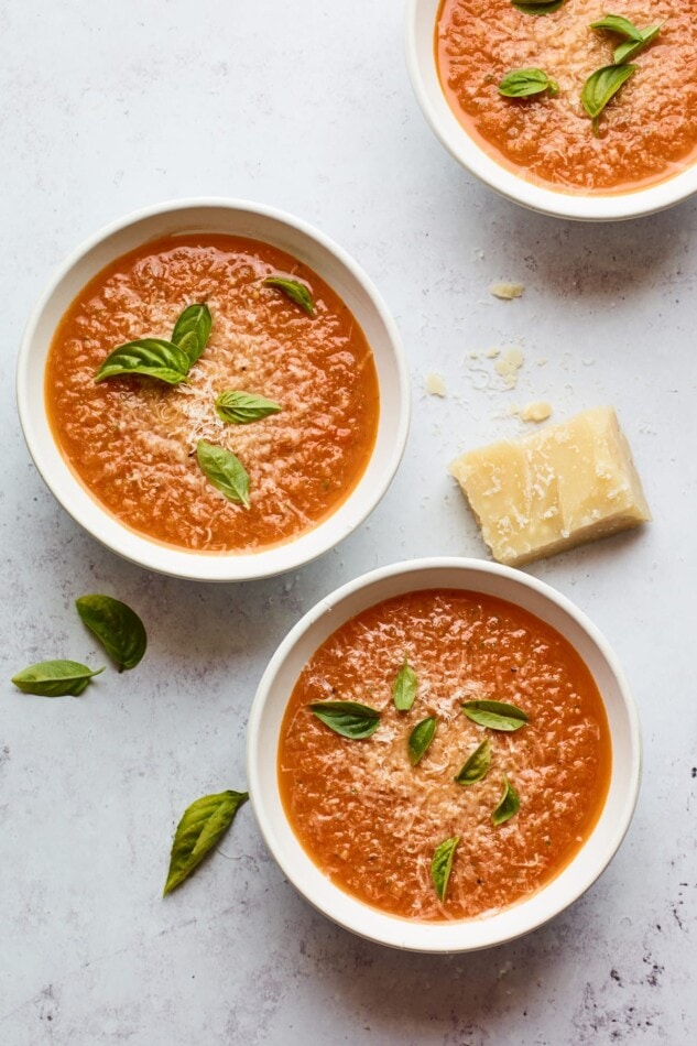Three bowls containing tomato basil soup. Each bowl is topped with fresh basil leaves and there is a wedge of parmesan cheese resting near the bowls.