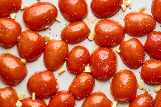 Garlic cloves and halved tomatoes coated in oil, salt and pepper spread across a baking sheet.