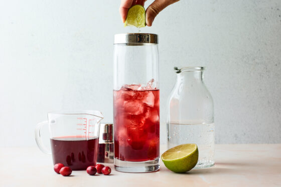 A clear shaker glass filled with vodka, cranberry and ice. A hand is squeezing fresh lime juice from a lime wedge into the shaker. Ingredients for vodka cranberry are scattered around the shaker.