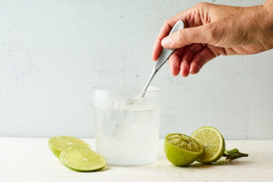 A hand mixing vodka, club soda and freshly squeezed lime in a drinking glass with a spoon.