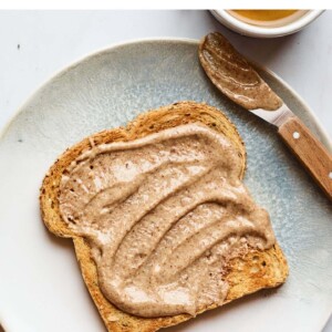 A slice of toast slathered with almond butter on a plate. There is a cup of tea next to the plate.