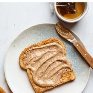 A slice of toast slathered with almond butter on a plate. There is a cup of tea next to the plate.