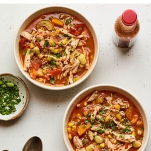 Three bowls of Brunswick stew. A bottle of hot sauce as well as a small bowl of fresh parsley are nearby for topping alongside two metal spoons.