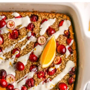 An overhead photo looking down at a baking dish containing cranberry orange baked oatmeal. There are whole and halved cranberries sprinkled on top next to orange slices and the oatmeal has been coated in a glaze.