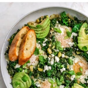 A skillet with green shakshuka that has been topped with slices of avocado and toasted bread.