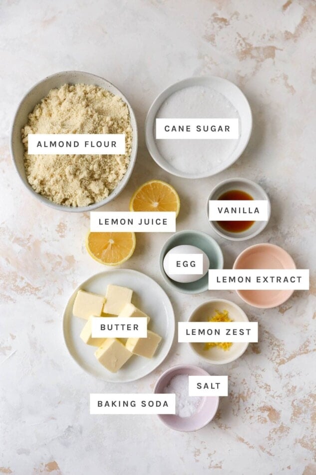 Ingredients measured out in bowls to make almond flour lemon cookies: almond flour, cane sugar, vanilla, lemon juice, egg, lemon extract, butter, lemon zest, salt and baking soda.