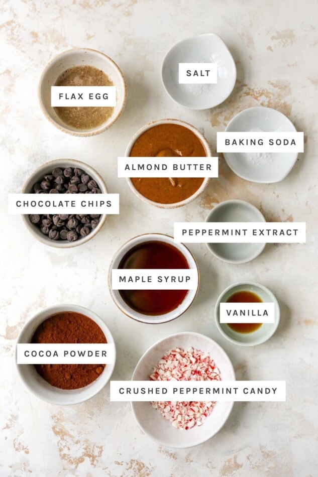 Ingredients measured out to make chocolate peppermint cookies: flax egg, salt, baking soda, almond butter, chocolate chips, peppermint extract, maple syrup, vanilla, cocoa powder and crushed peppermint candy.